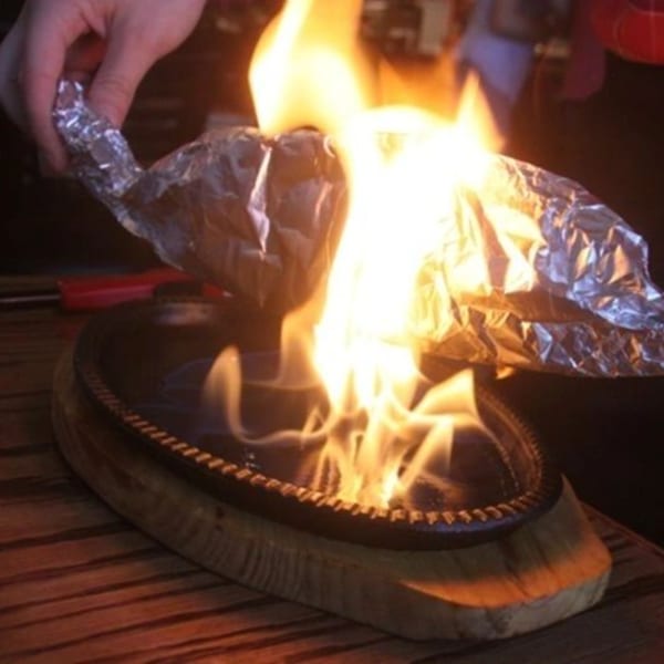 A hand holding foil-wrapped food over a sizzling hot plate with flames, creating a fiery presentation.