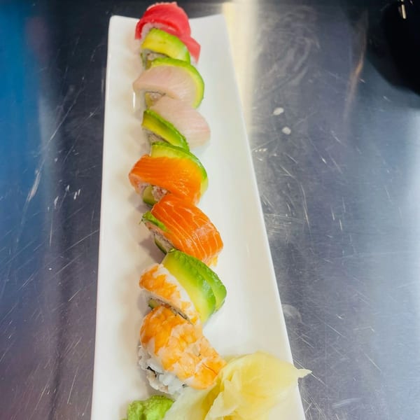 A long white plate holds a colorful sushi roll with salmon, tuna, shrimp, and avocado.